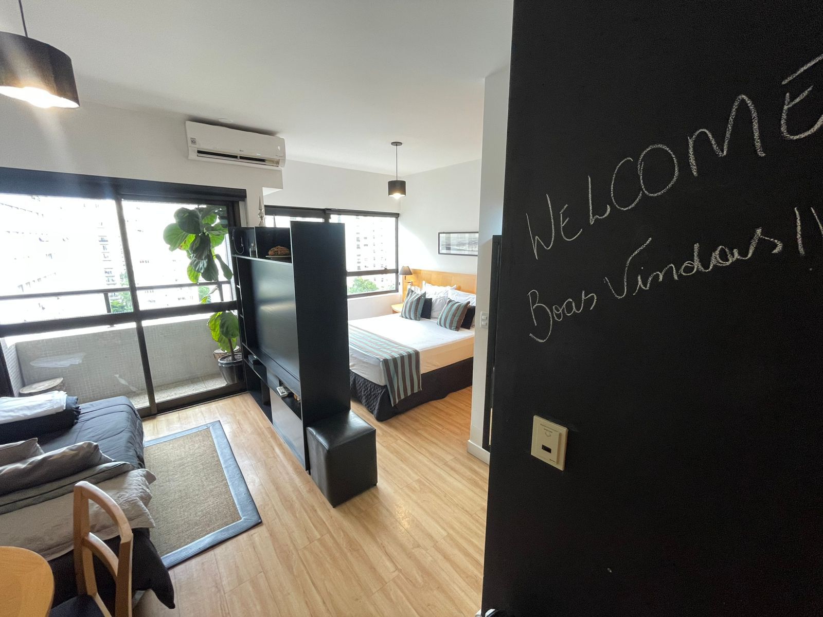 A modern hotel room with a large bed, a flat-screen TV, a sofa, and a desk. There is a blackboard with 'Welcome Boas Vindas!' written on it.