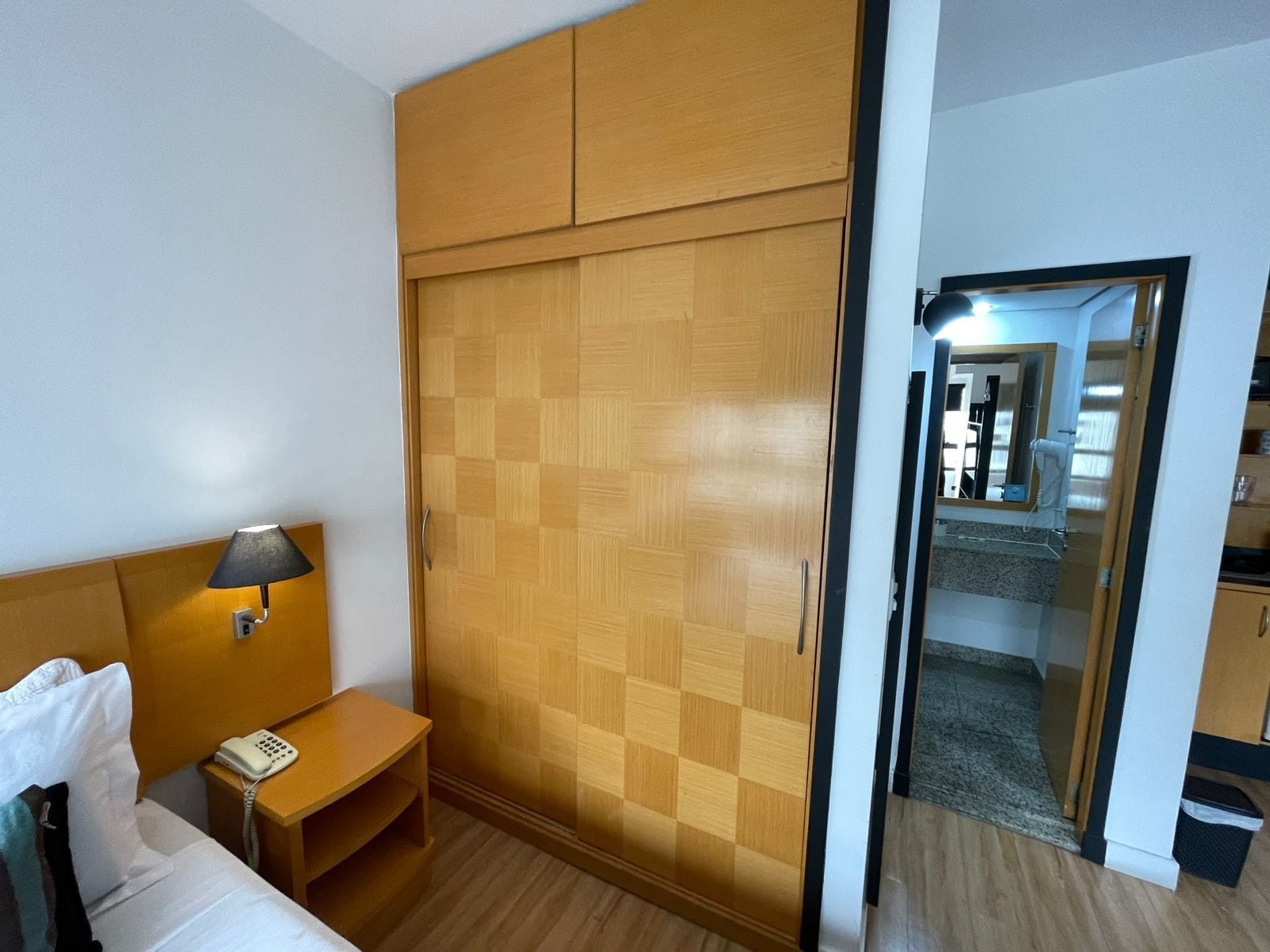 Quarto de hotel com cama, mesa de cabeceira, abajur e guarda-roupa xadrez.