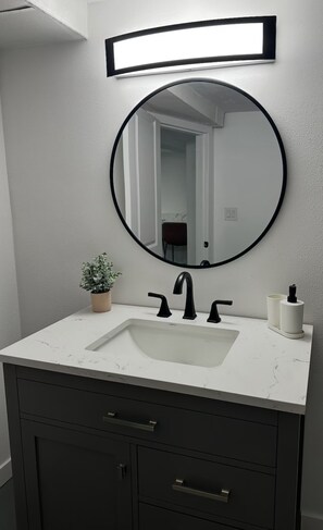 A bathroom with a white sink, black faucets, a round mirror, and a small potted plant. - The Little Harbor Retreat, Room Rental close to PSNS, Ferry, and more! (Bremerton)