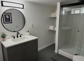 A modern bathroom with a round mirror, a sink, dark cabinets, a glass shower enclosure, and neatly stacked towels. - The Little Harbor Retreat, Room Rental close to PSNS, Ferry, and more! (Bremerton)