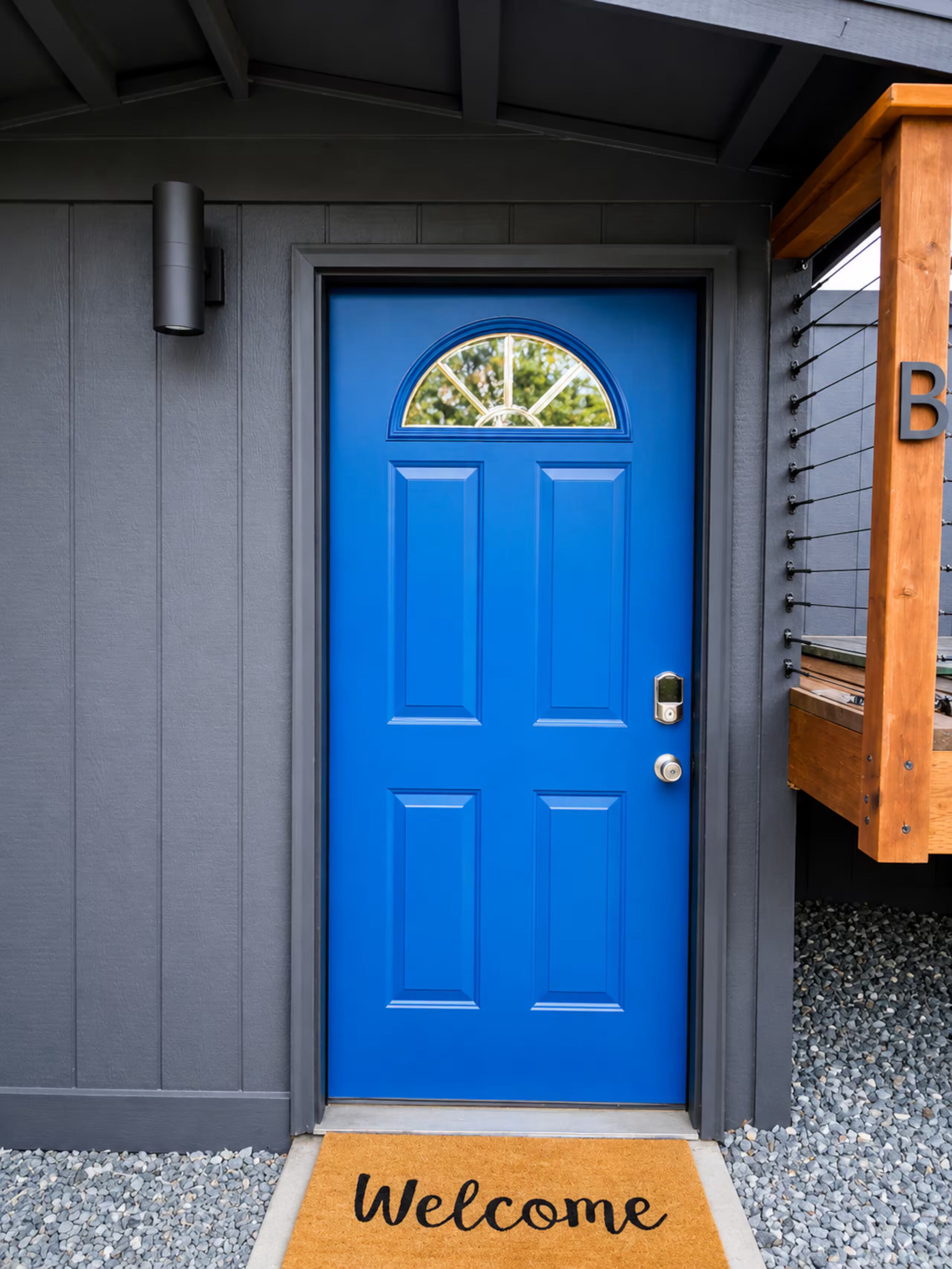 Une porte bleue avec un paillasson sur lequel est écrit « Bienvenue », une boîte aux lettres et une structure en bois portant la lettre « B ».