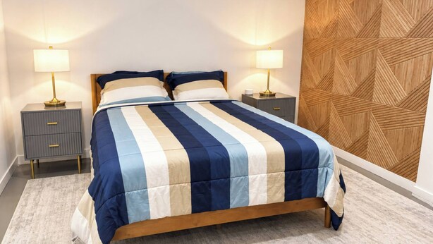 Ein Bett mit gestreifter Bettdecke, zwei Nachttische mit Lampen, ein hölzernes Kopfteil und eine Wand mit geometrischem Muster.