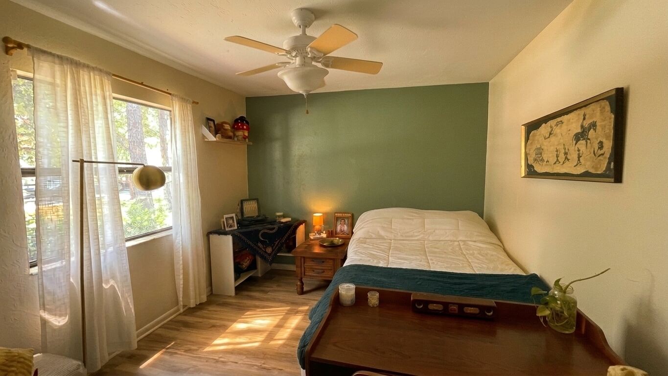 A bedroom with a bed, a ceiling fan, a window with curtains, a nightstand with a lamp, and a wall-mounted picture.