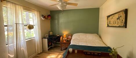 A bedroom with a bed, a ceiling fan, a window with curtains, a nightstand with a lamp, and a wall-mounted picture.