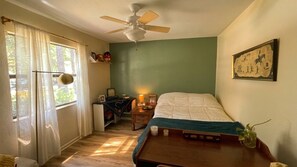 Une chambre à coucher avec un lit, un ventilateur de plafond, une fenêtre avec des rideaux, une table de chevet avec une lampe et un tableau accroché au mur.