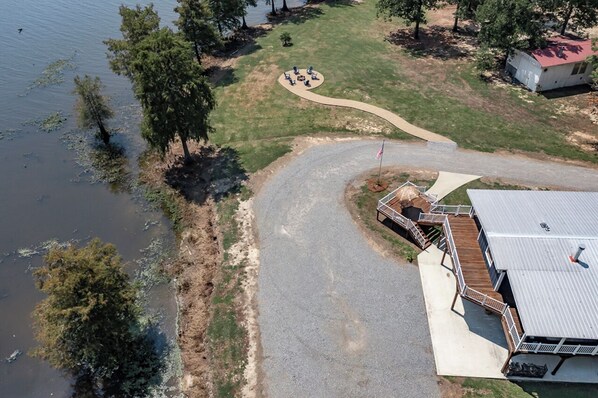 An aerial view of a lakeside property with a paved driveway, a dock, and a small building with a red roof. - Lakefront House with Outdoor Firepit, Grill, and More (Farmerville)