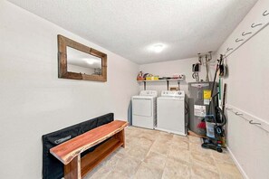 A laundry room with two washing machines, a bench, a mirror, and a vacuum cleaner. - Salted Skyline | Private Community Beach Access | Hot Tub and Dog Friendly (Kitty Hawk)