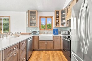 A modern kitchen with wooden cabinets, stainless steel appliances, and a large window. - Salted Skyline | Private Community Beach Access | Hot Tub and Dog Friendly (Kitty Hawk)