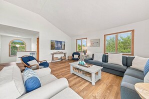 A spacious living room with a large sofa, a coffee table, and a dining table. There are two windows with blinds, a door, and a wall-mounted artwork. - Salted Skyline | Private Community Beach Access | Hot Tub and Dog Friendly (Kitty Hawk)
