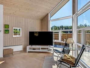 A modern living room with a large flat-screen TV, wooden panel walls, a glass table with chairs, and a view of trees through large windows. - "Isbiorn" - 800m from the sea by Interhome (Løkken)