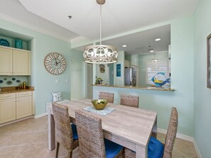 A dining area with a wooden table, wicker chairs, a chandelier, and a kitchen in the background. - Marco Island 2 king bed unit + pool with park views. Walk to shops & dining. (Marco Island)