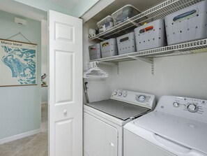 A laundry room with a washing machine, dryer, and shelves with storage bins. - Marco Island 2 king bed unit + pool with park views. Walk to shops & dining. (Marco Island)