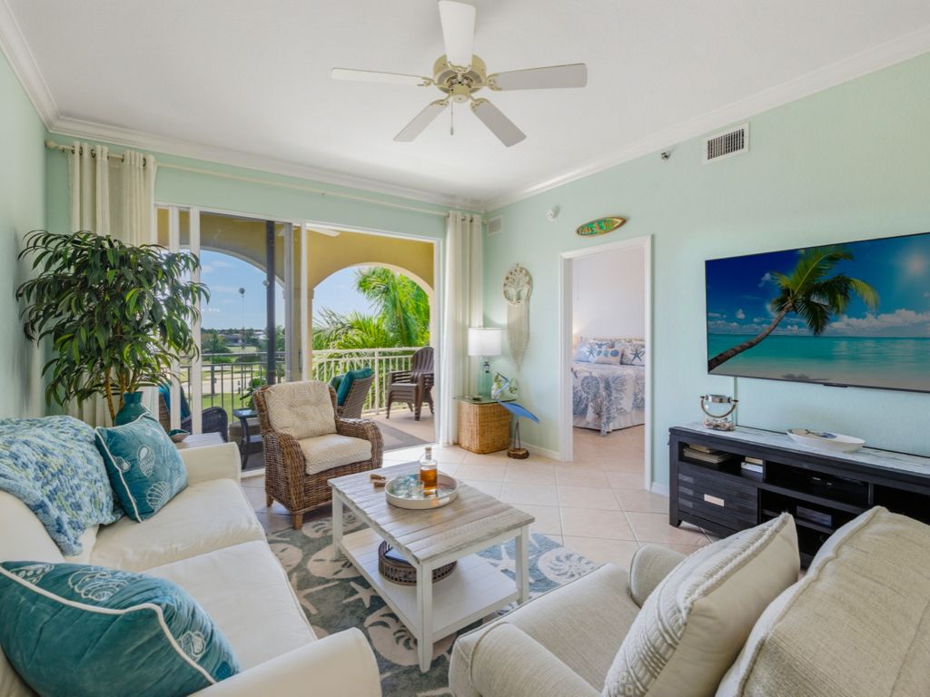 A living room with a sofa, coffee table, and television. There is a balcony with a view of palm trees.