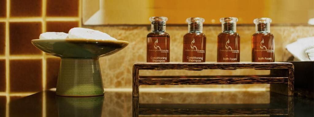 A bathroom with a tiled wall, a green vase with a white object, and four bottles labeled 'Conditioning Shampoo' and 'Bath Foam' on a wooden shelf.