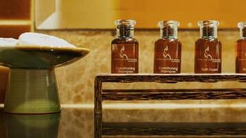 A bathroom with a tiled wall, a green vase with a white object, and four bottles labeled 'Conditioning Shampoo' and 'Bath Foam' on a wooden shelf.