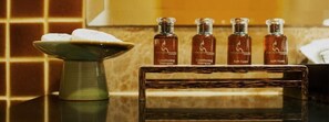 Un baño con pared de azulejos, un jarrón verde con un objeto blanco, y cuatro botellas etiquetadas como 'Conditioning Shampoo' y 'Bath Foam' sobre un estante de madera.