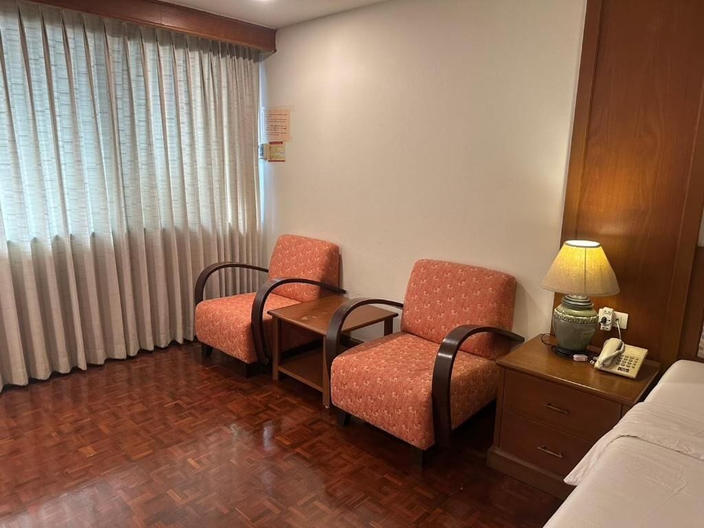 Habitación de hotel con dos sillones, una mesita de noche de madera con lámpara, teléfono y cama.