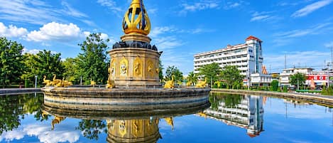 En springvand med en central konstruktion og omgivende statuer, beliggende i et spejlende vandareal, med en bygning i baggrunden.