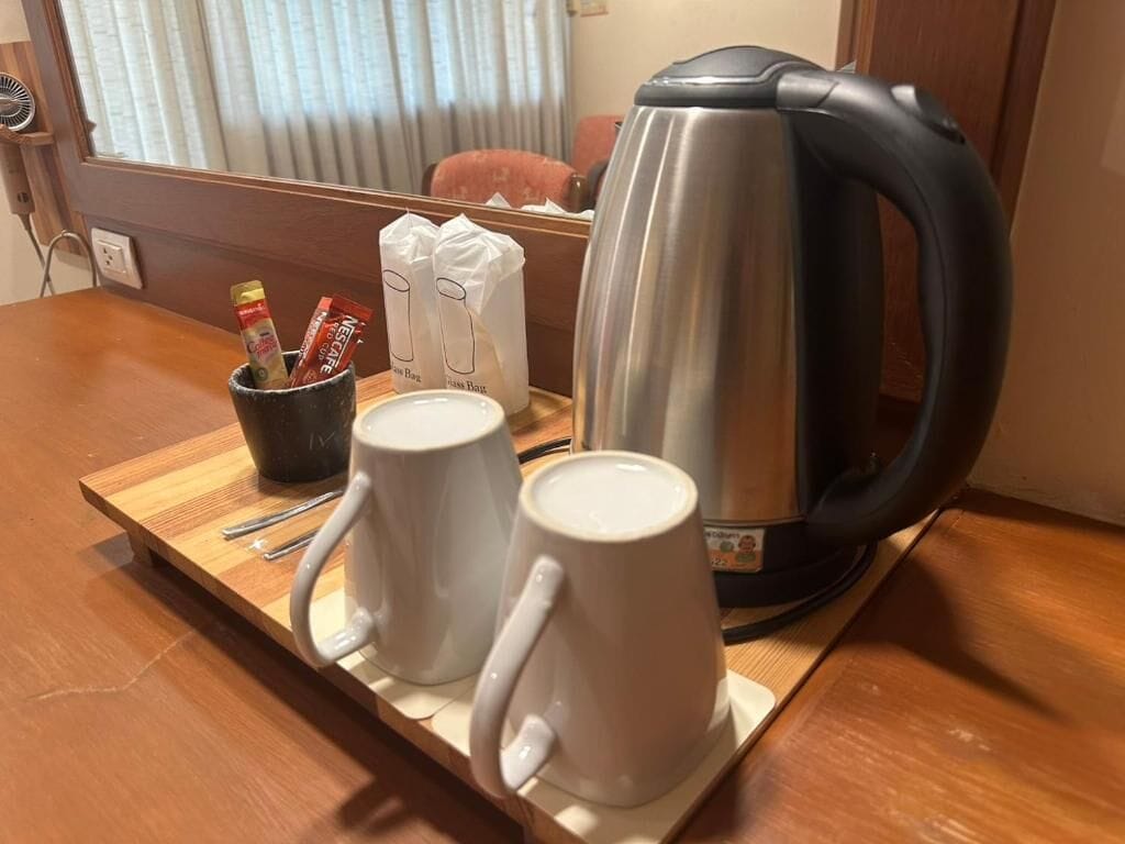 A wooden table with two white mugs, a stainless steel kettle, and a black mug with a handle.