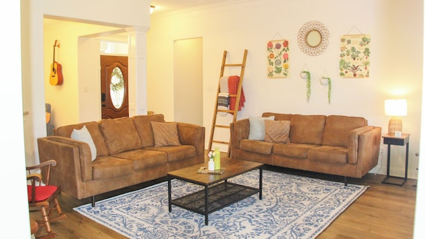 A living room with two brown sofas, a coffee table, a patterned rug, a guitar, a ladder, and wall decorations including a floral print and a mirror. - Aryn Ridge House / 3BR / Near Casinos & Interstate (Lake Charles)