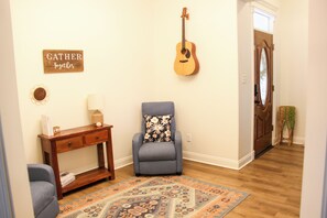 A living room with a grey armchair, a wooden console table, a guitar, a rug, and a door with a mirror.