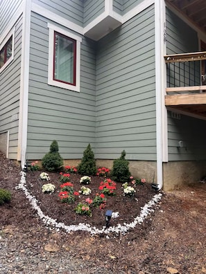 Une maison avec une fenêtre d’angle, une terrasse et un parterre fleuri avec des fleurs rouges et blanches.