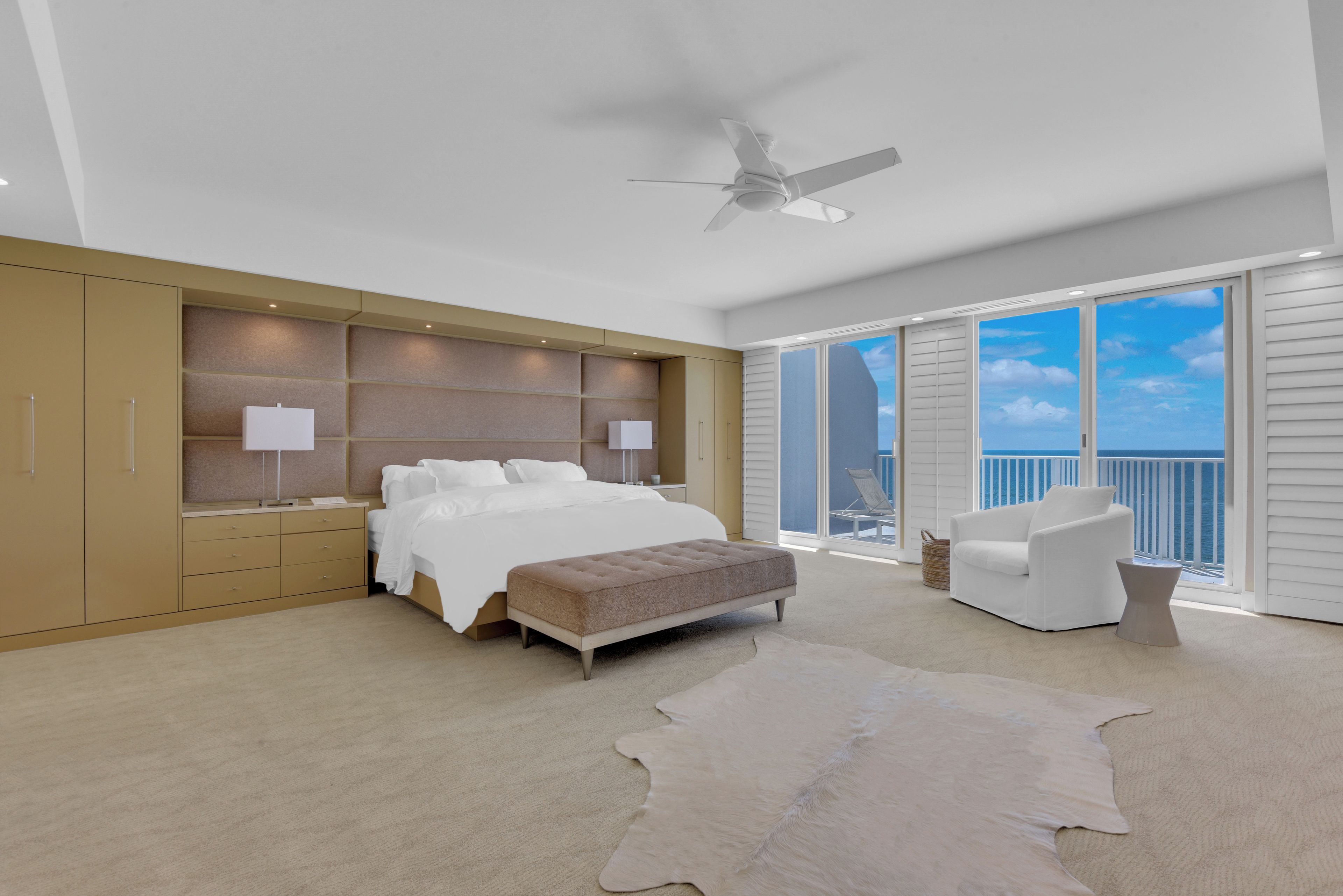 A bedroom with a bed, bedside tables, a ceiling fan, a large window with a view, and a balcony with a chair.