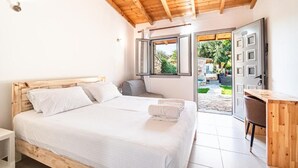 Un dormitorio con techo de madera, una cama con sábanas blancas, un escritorio pequeño y una puerta abierta que da a un jardín.