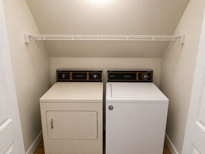 A laundry room with two stacked washers and dryers, a metal shelf above, and a white door on the left. - Pecker’s Nest  Cozy Cottage in Galena Territory (Galena)