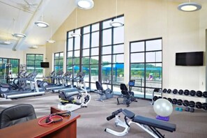 Academia com diversos equipamentos de ginástica, amplas janelas e vista para o verde.