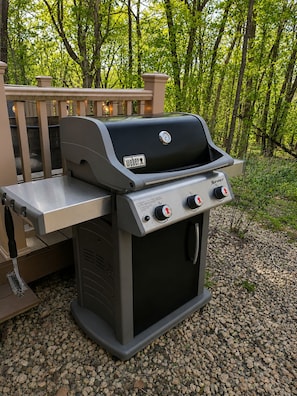 Una parrilla Weber Spirit portátil con una mesita auxiliar, sobre una superficie de grava con una terraza de madera y follaje de fondo.