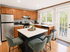 A modern kitchen with wooden cabinets, stainless steel appliances, a central island with bar stools, and a view through a glass door. - Pecker’s Nest  Cozy Cottage in Galena Territory (Galena)