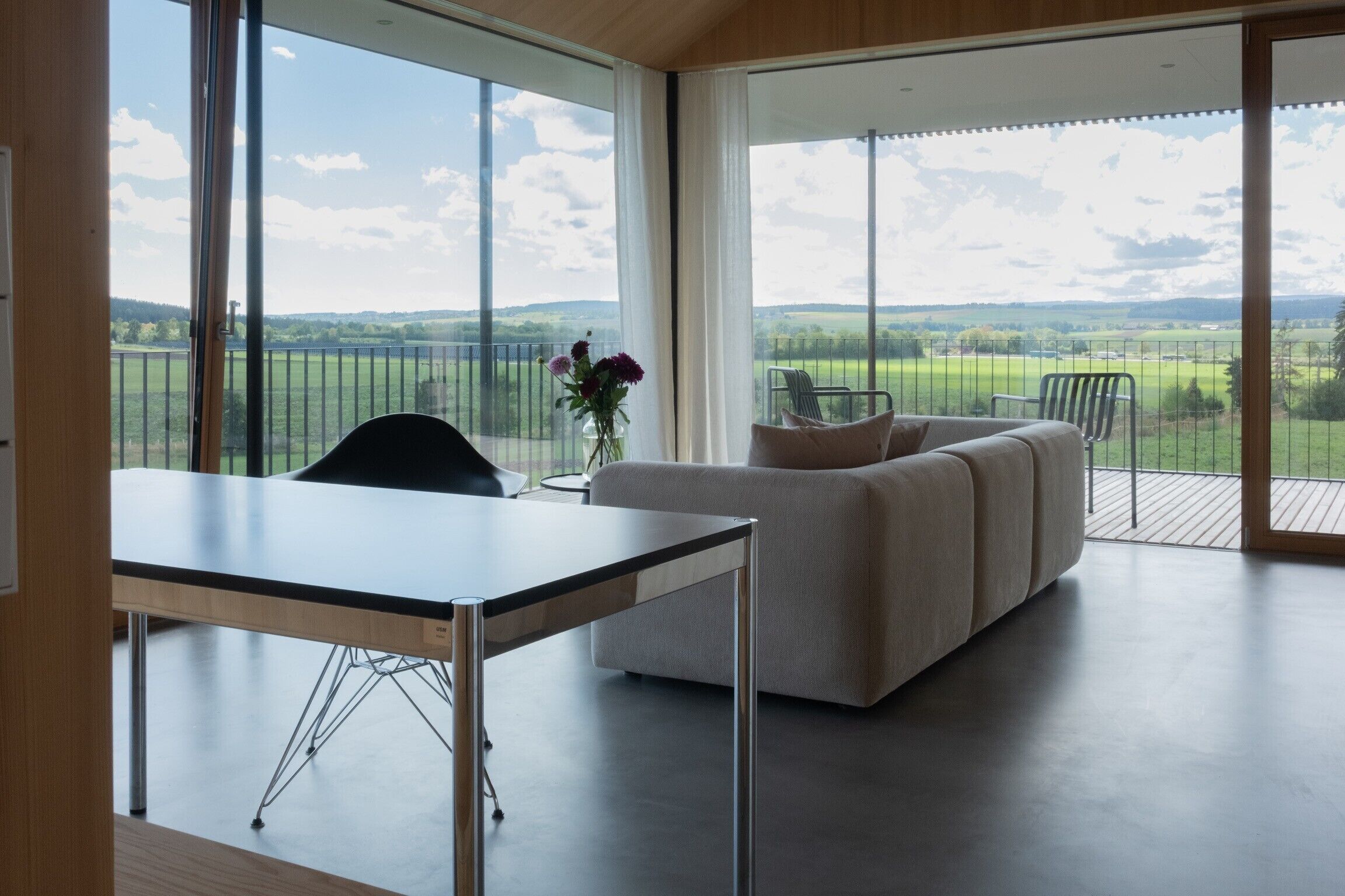 A modern interior with a dining table, a sofa, and a view of a green landscape through large windows.