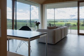 Ein modernes Interieur mit einem Esstisch, einer Couch und Blick auf eine grüne Landschaft durch große Fenster.