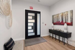 A modern interior with a black-framed door marked 'EXIT,' a black console table with two white water bottles, a woven basket, and a large forest-themed painting on the wall. - Desert Sands Basecamp | Hot Tub | Pool | Fire Pit (Hurricane)
