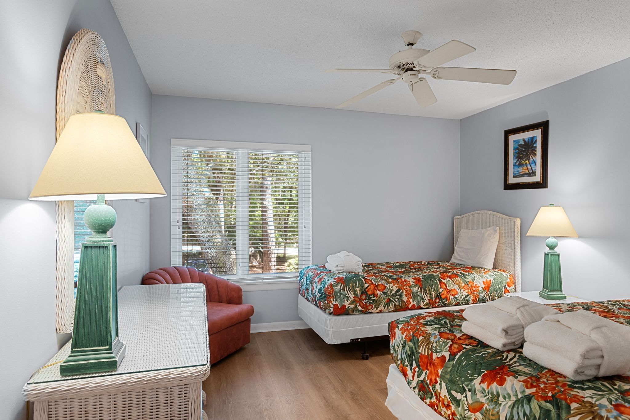 A bedroom with a bed, two bedside tables, a red armchair, a ceiling fan, and a window with blinds.