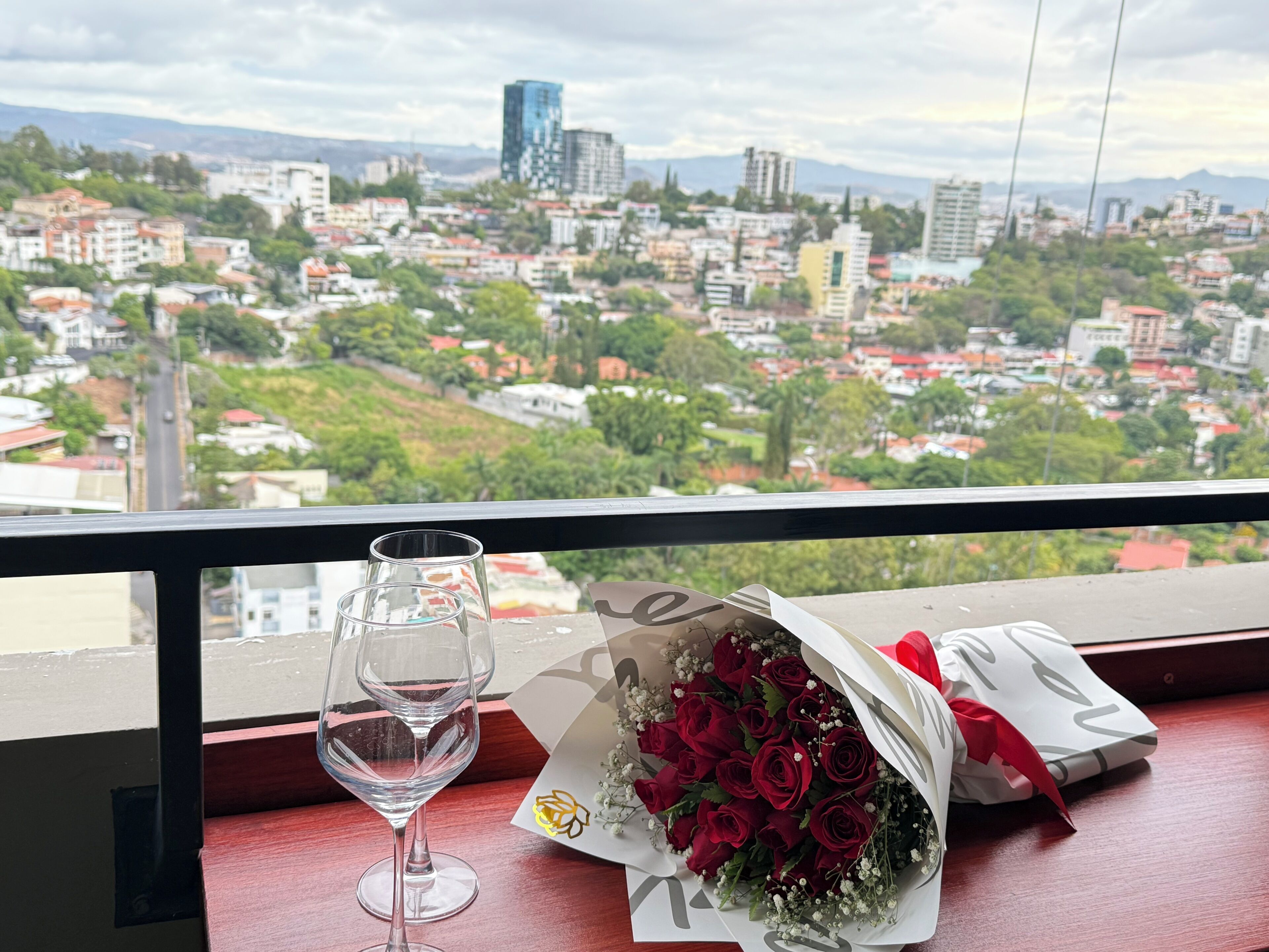 A balcony with a view of a cityscape, a glass of wine, and a bouquet of red roses.