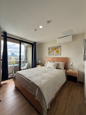 Een hotelkamer met een groot bed, een nachtkastje met een lamp, uitzicht op de stad door het raam en een airconditioning aan de muur.