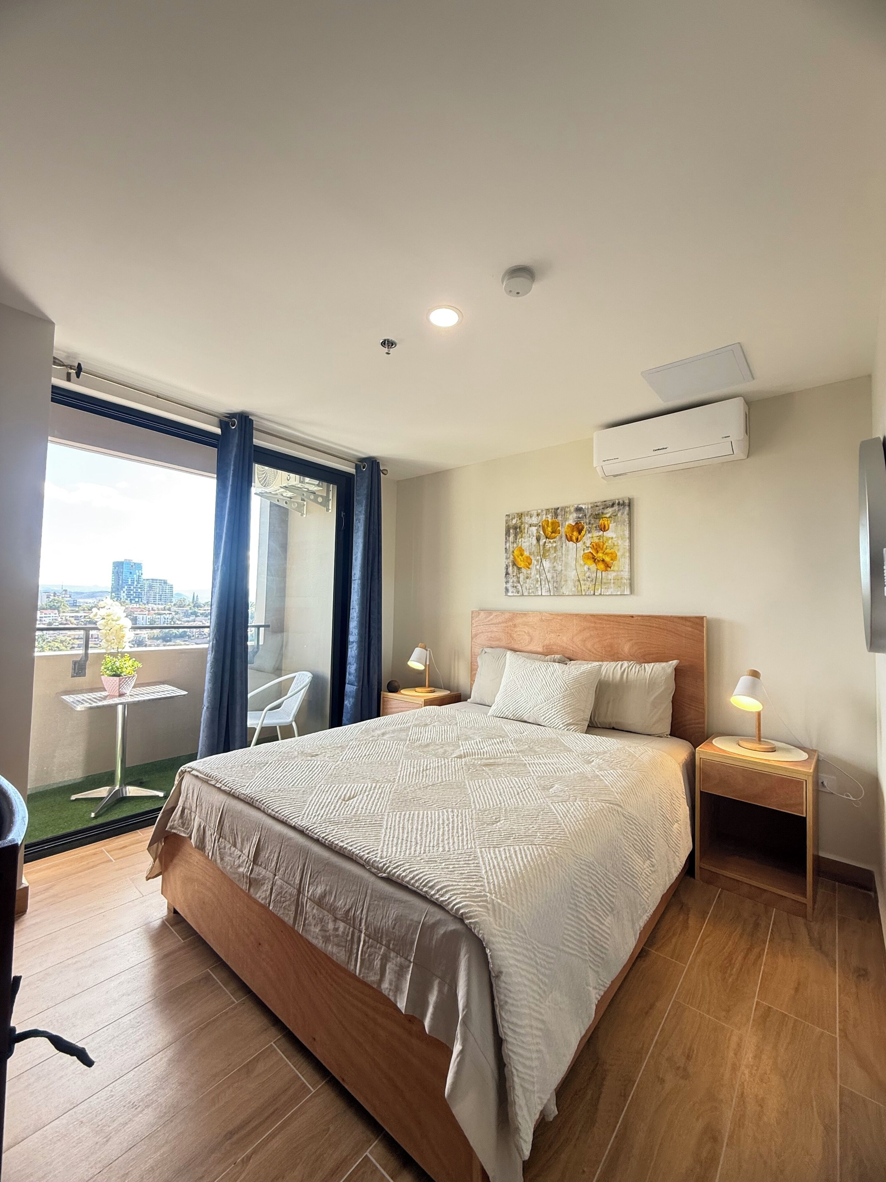 Habitación de hotel con una cama grande, una mesita de noche con lámpara, una pequeña mesa con planta en maceta, vista de la ciudad desde un balcón y un equipo de aire acondicionado en la pared.