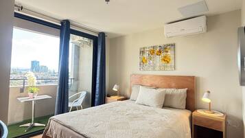 Habitación de hotel con una cama grande, una mesita de noche con lámpara, una pequeña mesa con planta en maceta, vista de la ciudad desde un balcón y un equipo de aire acondicionado en la pared.