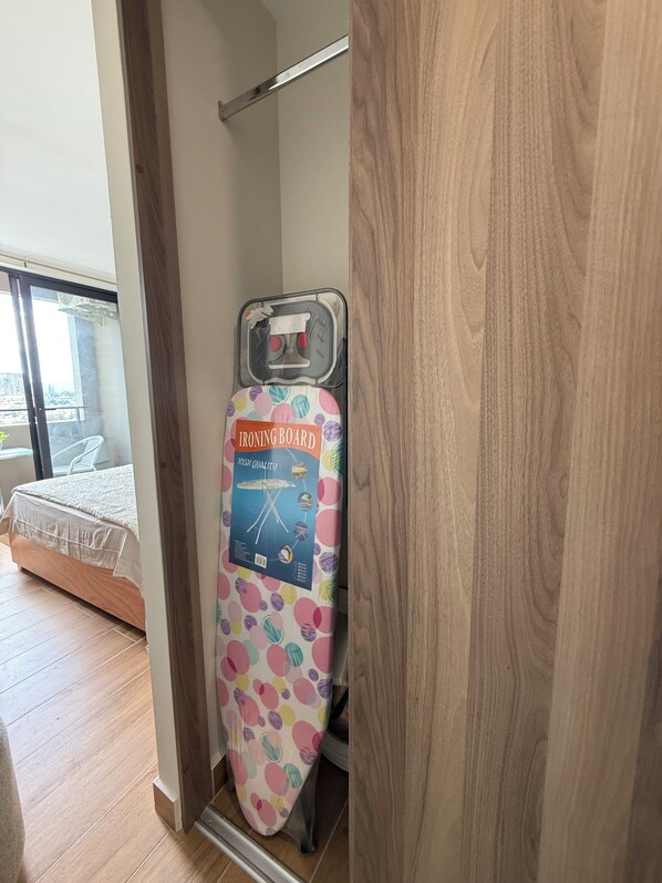 Een kamer met een bed, een houten vloer, een raam met uitzicht en een strijkplank met een kleurrijke stippenhoes.
