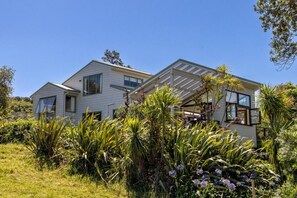 A two-story house with a covered patio area, surrounded by lush greenery and flowering plants. - Coastal Calm by Waiheke Pure (Waiheke Island)
