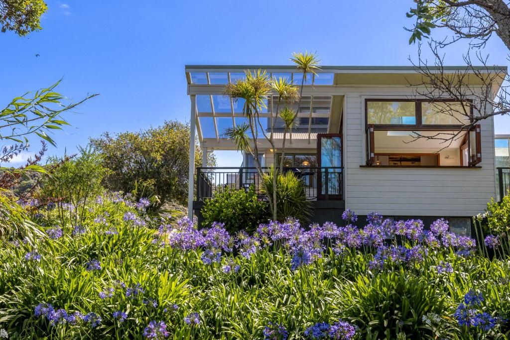 A modern two-story house with a glass facade, surrounded by a garden with purple flowers.