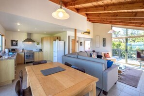 A modern kitchen with wooden beams, a dining area with a table, and a living space with a sofa. - Coastal Calm by Waiheke Pure (Waiheke Island)