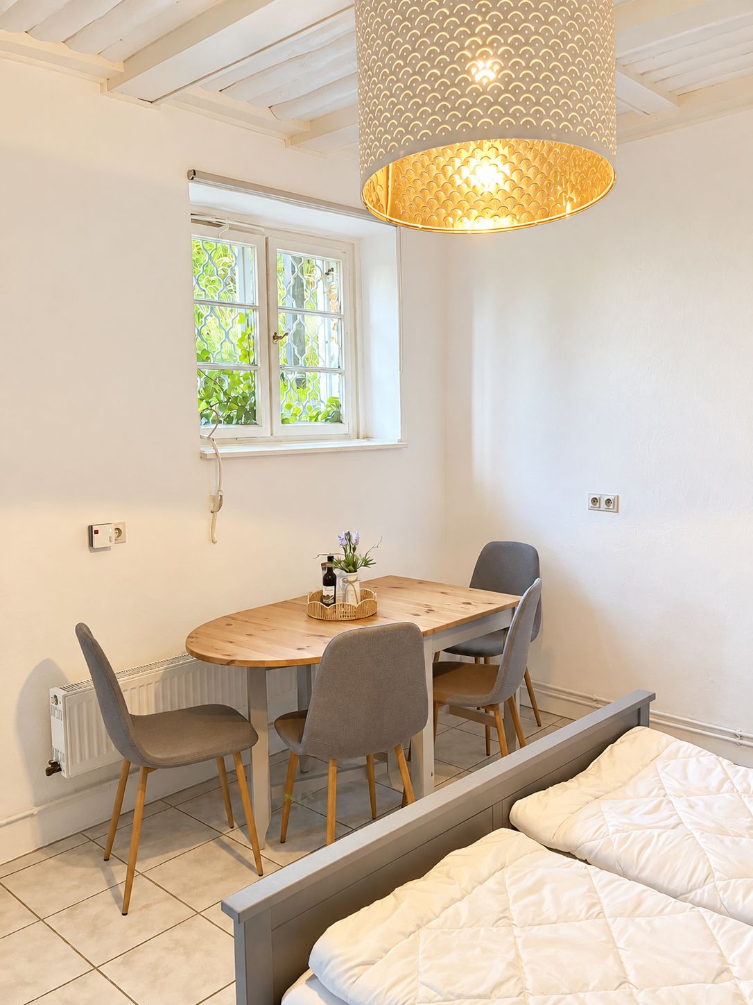 Een kamer met een bed, een eettafel met stoelen, een hanglamp en een raam met uitzicht op groen.