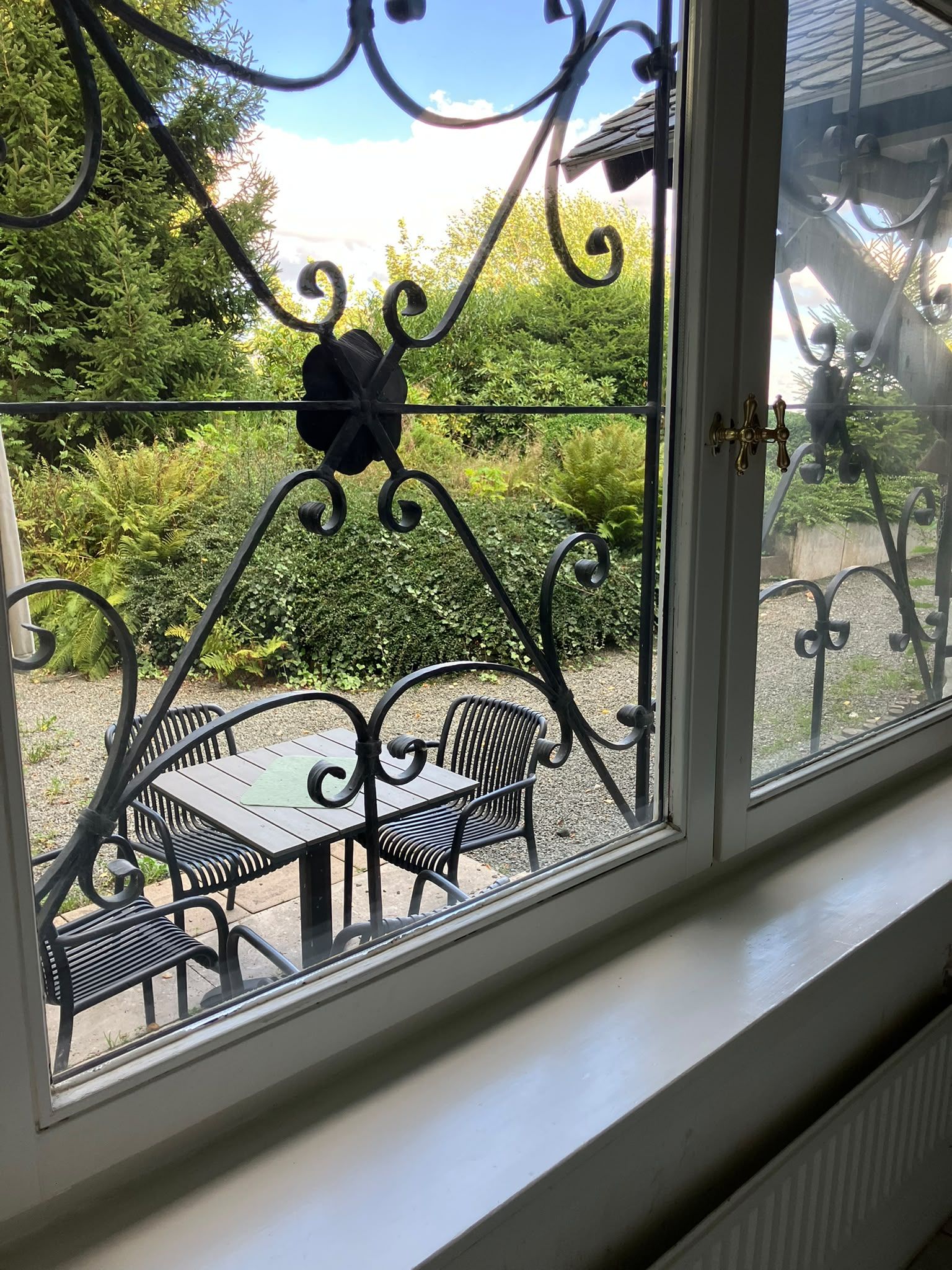 Ein Fenster mit Blick auf eine Außenpatio-Terrasse, auf der sich ein Tisch und Stühle befinden, umgeben von viel Grün.