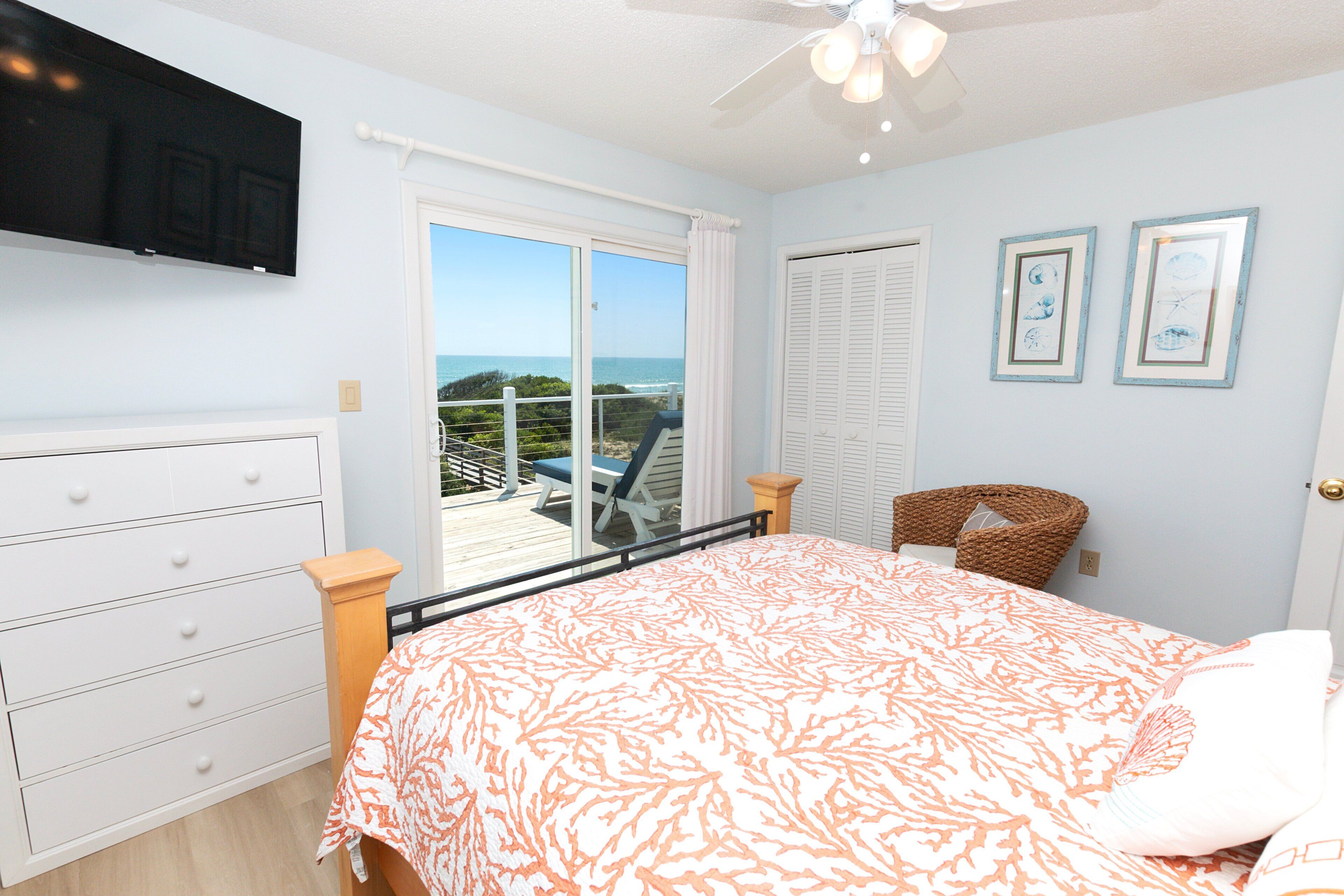 A bedroom with a bed, a dresser, a chair, a television, and a view of the ocean through a sliding door.
