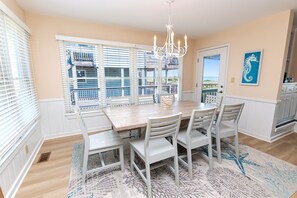 A dining area with a wooden table, white chairs, a chandelier, and a view of a balcony with a railing. - OCEANFRONT, Ocean Views, Hot Tub, Comm Pool -104, Shore Crew (Corolla)