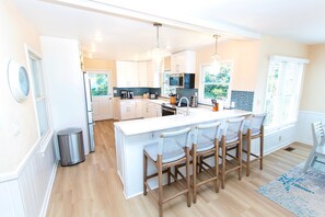 A modern kitchen with white cabinetry, a central island with bar stools, and a large window providing natural light. - OCEANFRONT, Ocean Views, Hot Tub, Comm Pool -104, Shore Crew (Corolla)