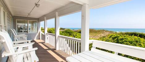 A covered porch with white railings and benches, overlooking a beach with vegetation and the ocean.